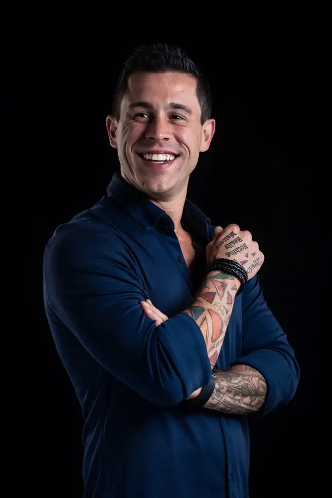 Johnny Destro de braços cruzados, camisa azul-marinho, sorrindo, sobre fundo preto